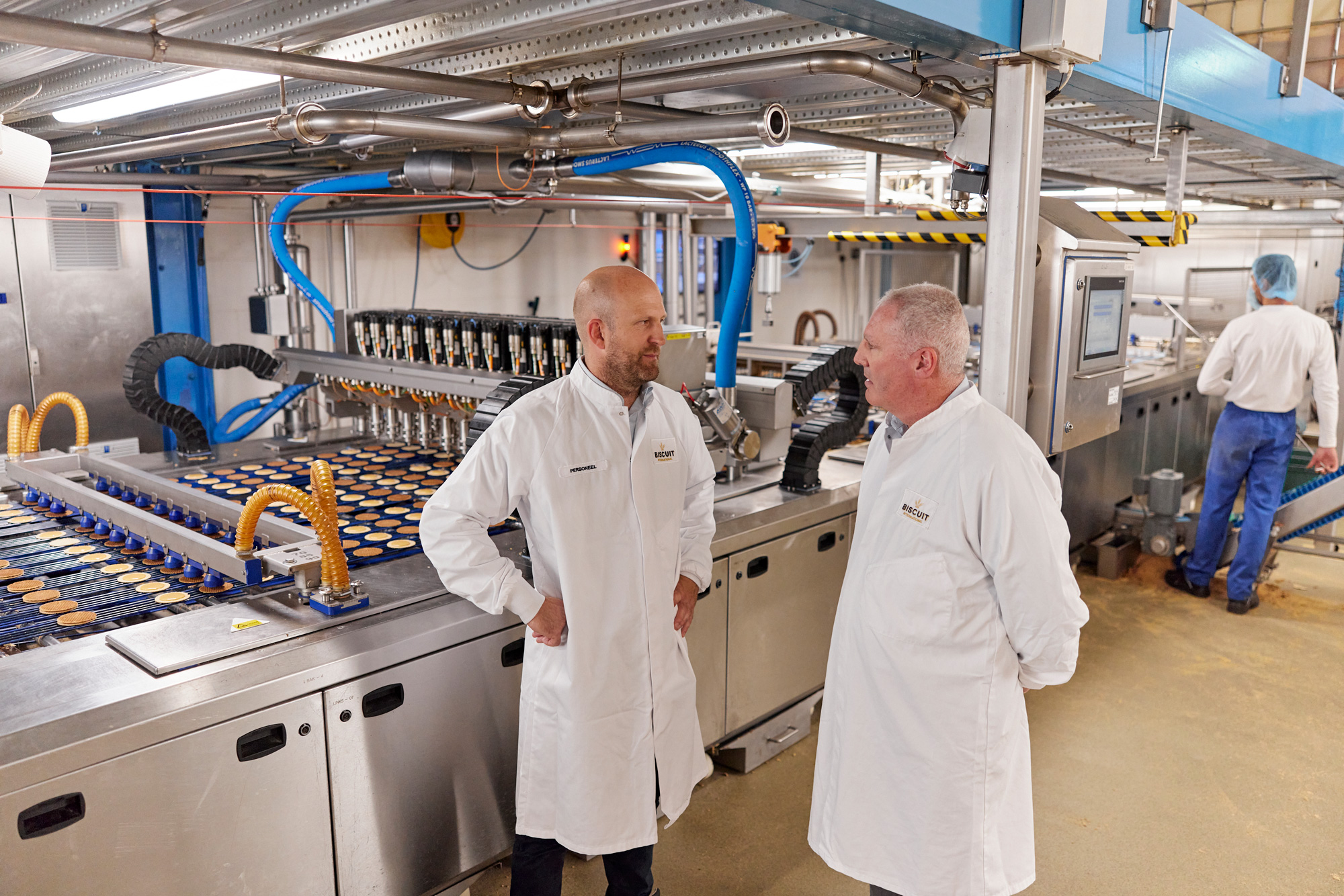 Two men in lab coats talking in an industrial food processing plant with conveyors and packaged products moving in the background.