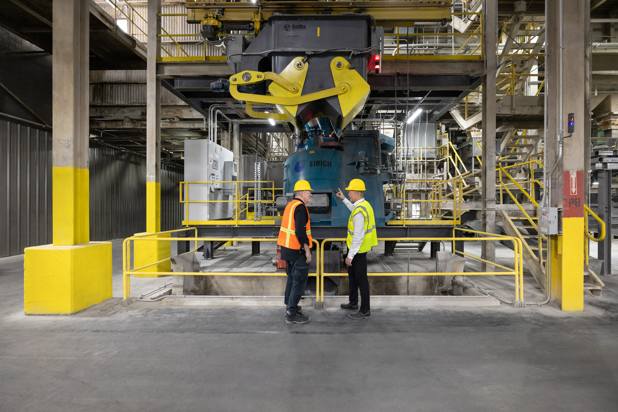 Two workers in hard hats and safety vests inspect machinery in an industrial setting with large yellow robotic arms and steel structures.