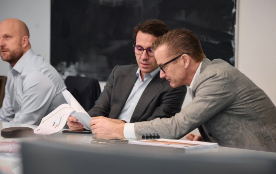 Two men in suits reviewing documents together at a conference table while another man listens, in a modern office setting.