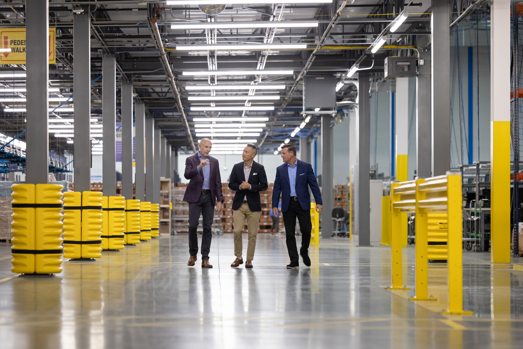 Three business professionals, two men and one woman, walk through a large industrial warehouse with yellow containers and structural columns, engaged in a conversation.