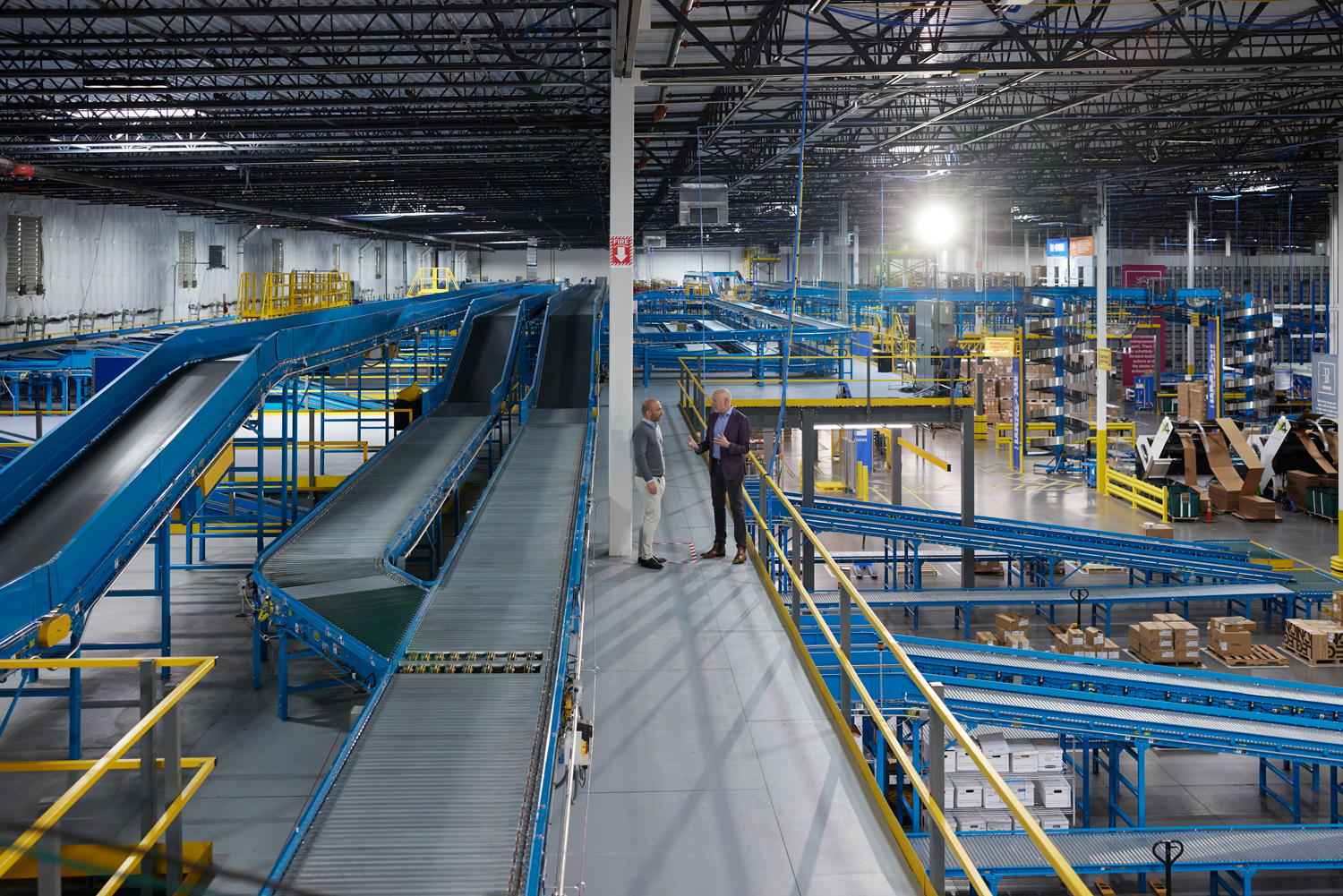 The image shows 2 man standing in a warehouse. The setting includes steel structures, stairs, and an industrial atmosphere.