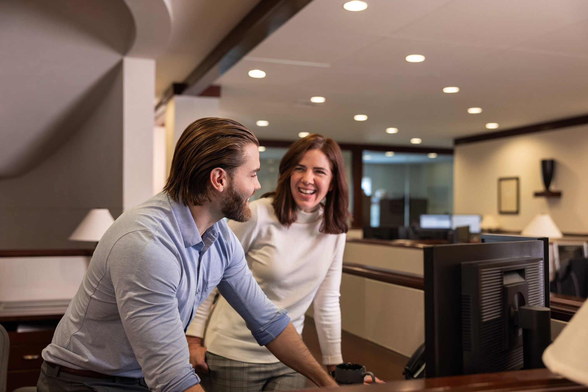 Two colleagues, a man and a woman, happily interact at an office desk, suggesting a friendly conversation in a professional setting.