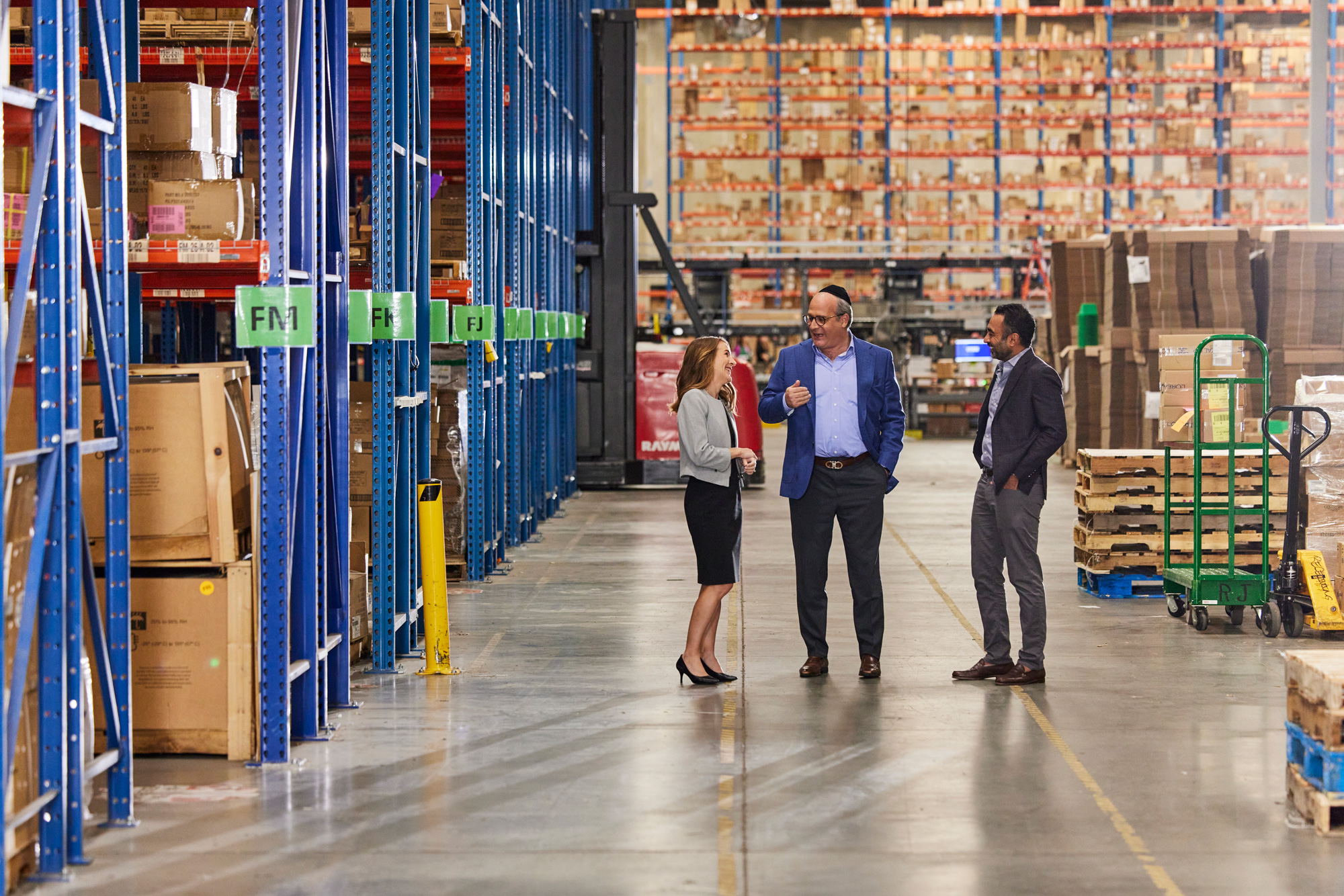 Three professionals converse while walking in a spacious warehouse with tall shelving stocked with various goods.