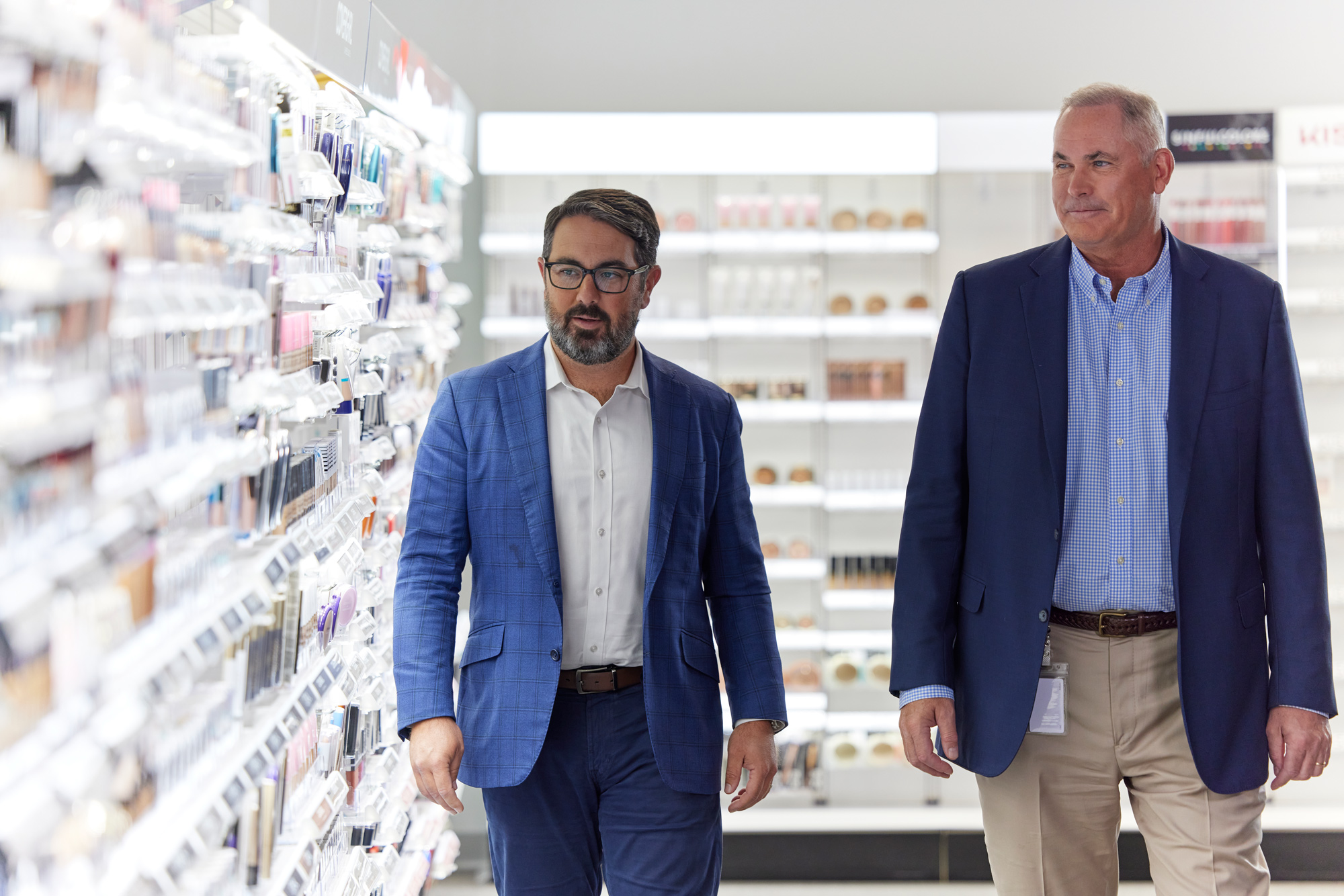 Two businessmen walking through a store aisle with shelves stocked with products, focusing intently as they discuss.